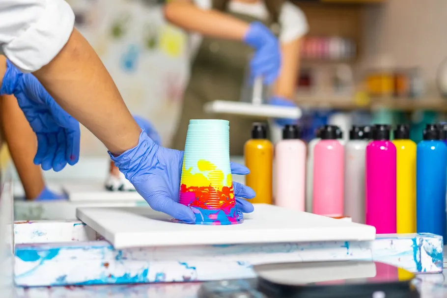 Gloved hands paint-pouring onto canvas.