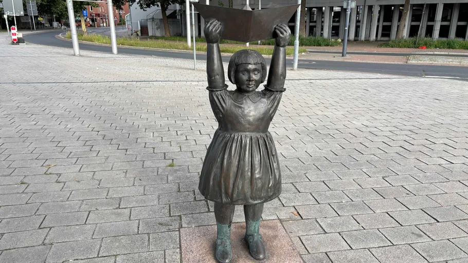 Bronze girl statue holding paper boat outdoors.