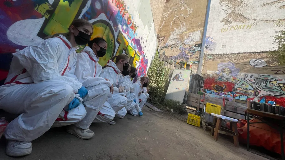 People in white suits crouching by a graffiti wall.