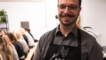 Mann lächelt mit Schürze, Menschen sitzen dahinter.