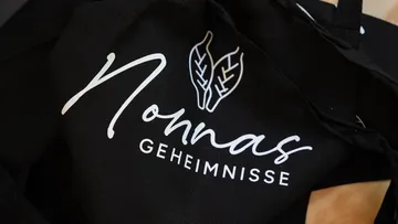 Schwarze Schürze mit 'Nonnas Geheimnisse' und Blättern.