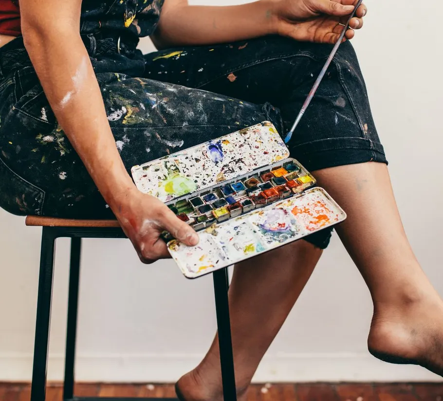 Person holding watercolor palette, seated on stool.