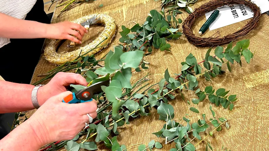 Hands crafting wreaths with greenery on table.