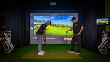 Two people practicing golf indoors with simulator.