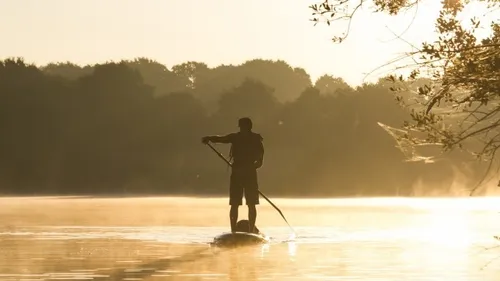 Person beim Stand-up-Paddeln auf ruhigem See bei Sonnenaufgang.