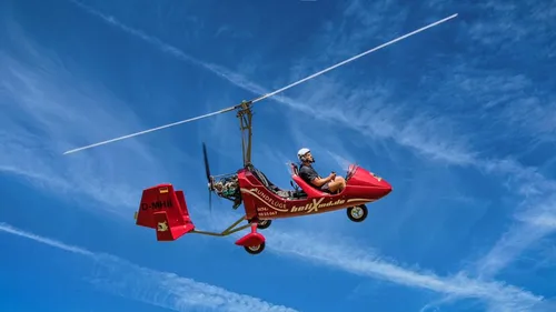 Red autogyro flying under clear blue sky.