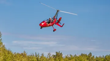Red gyrocopter flying over a forested area.