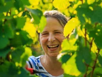Frau lächelt zwischen grünen Weinblättern im Sonnenschein.