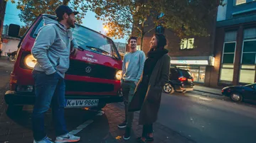Three people talking beside red van on street.