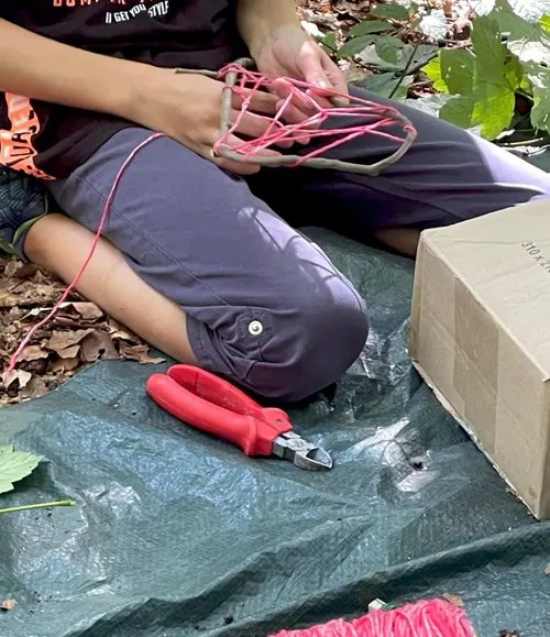 Person weaving pink thread on sticks outdoors.