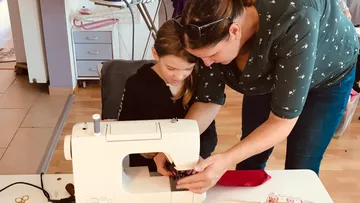Woman teaching girl to sew with machine indoors.