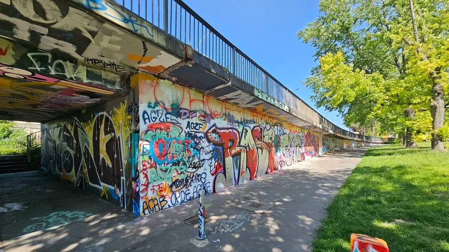 Graffiti-bedeckter Gehweg unter Brücke, sonniger Park daneben.