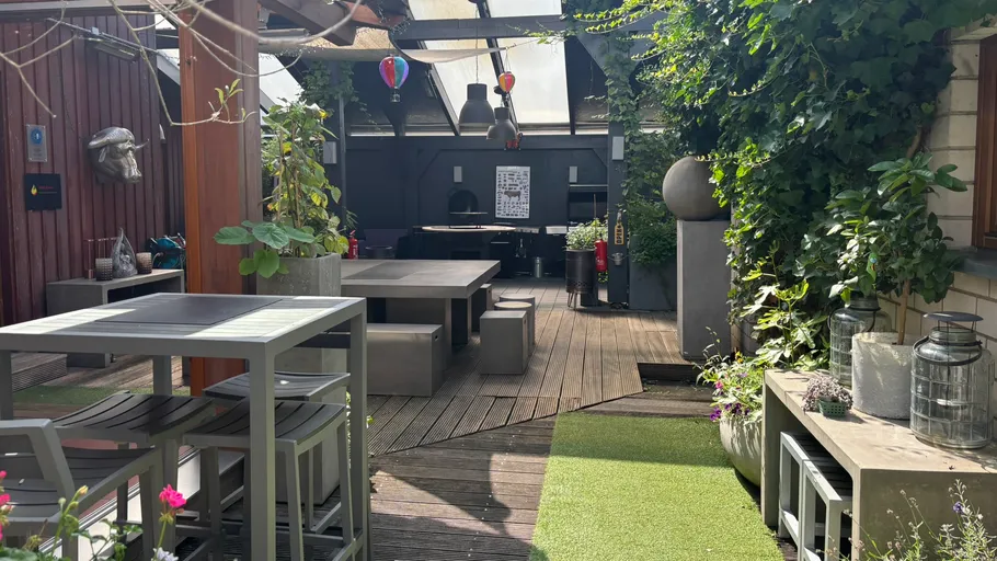 Überdachte Terrasse mit Tischen, Pflanzen und Sitzgelegenheiten.