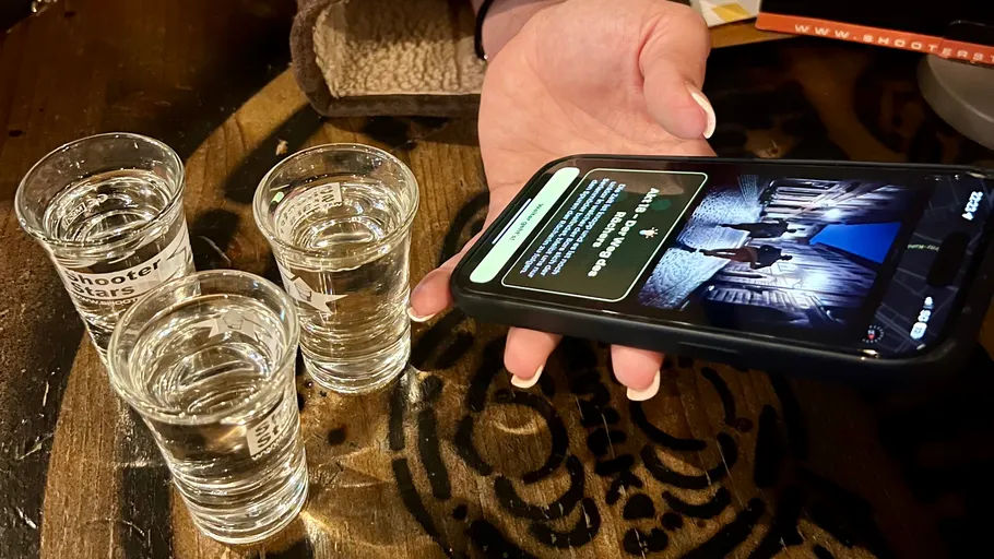 Hand hält Telefon neben drei Schnapsgläsern.