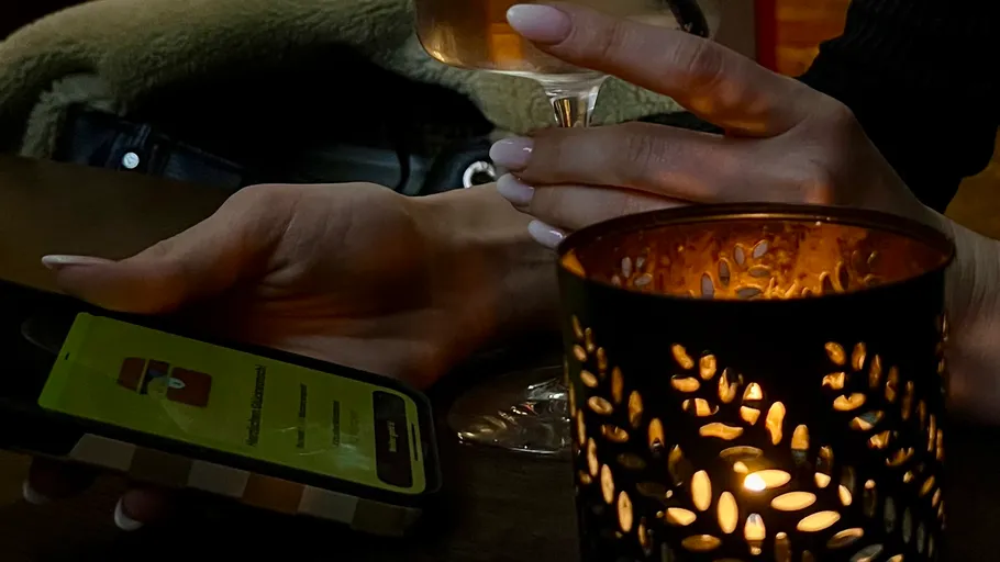 Hand hält Telefon und Weinglas, Kerze nahebei.