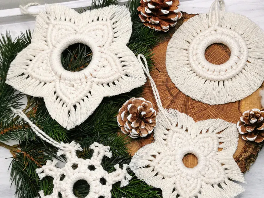 Weiße Makramee-Ornamente liegen auf Tannenzweigen und Holz.