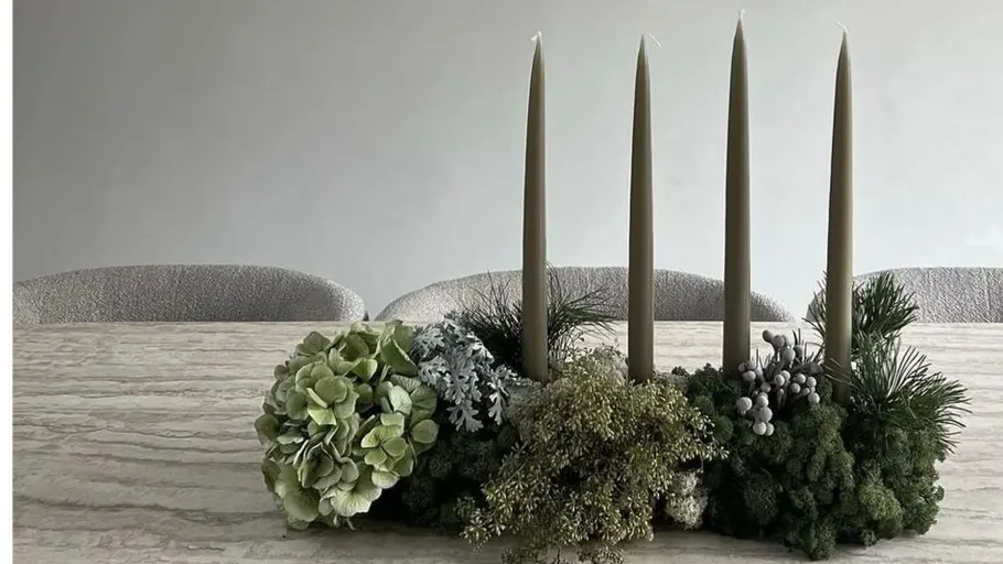 Vier Kerzen in Blumengesteck auf Tisch.