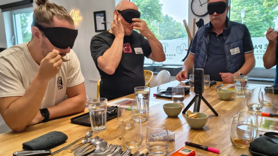 Personen mit Augenbinden probieren Essen an einem Tisch.