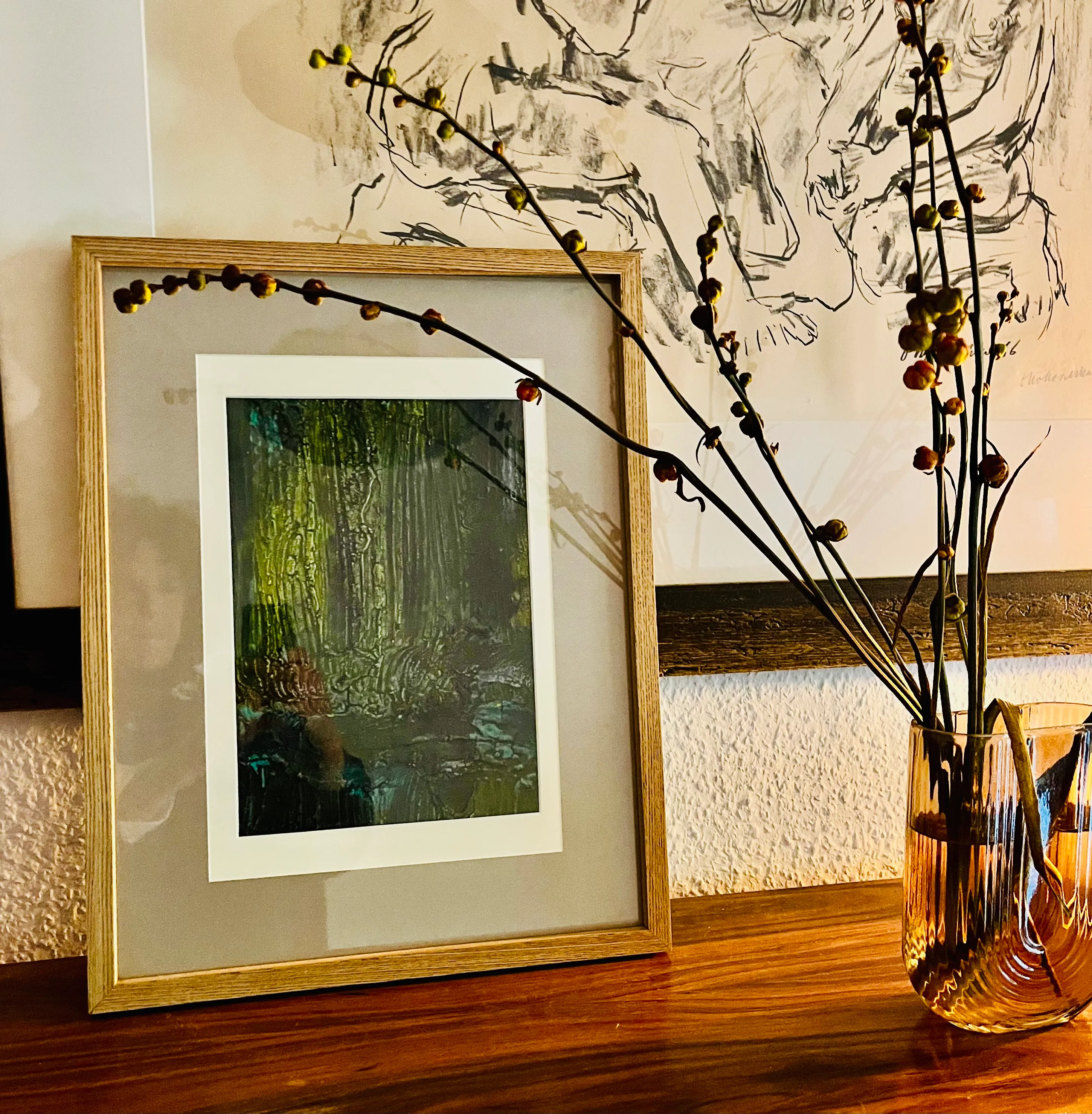Gerahmtes Kunstwerk neben Vase mit Zweigen auf Tisch.