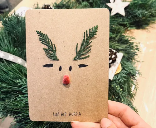 Hand holding reindeer-themed card with greenery background.