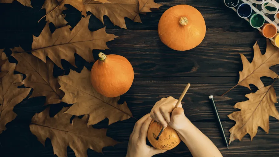 Person drawing on pumpkin surrounded by leaves.