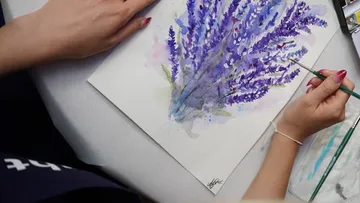 Person painting purple flowers on canvas.