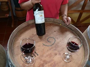 Eine Flasche Wein steht auf einem Holzfass, zwei Gläser daneben.