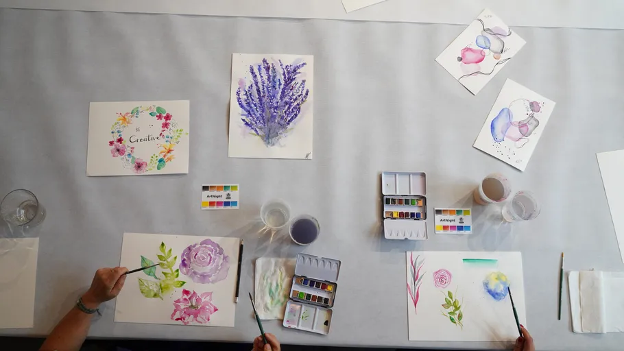 Zwei Hände malen Blumen mit Wasserfarben auf Tisch.