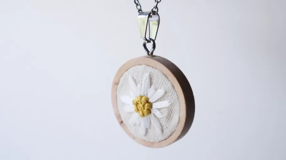 Embroidered flower pendant on chain, minimalistic backdrop.