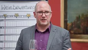 Mann hält Weinglas, spricht, mit Tafel dahinter.