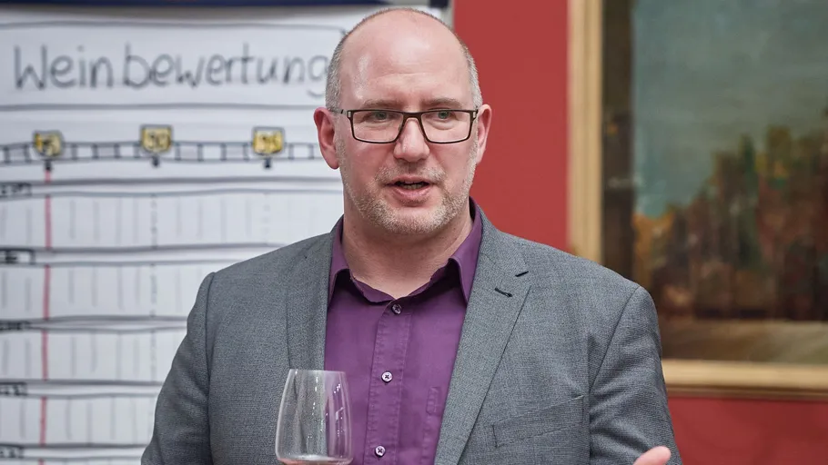 Mann hält Weinglas, spricht, mit Tafel dahinter.