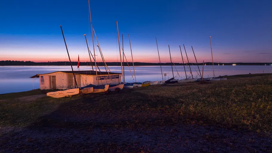 Segelboote und Schuppen an ruhigem See bei Dämmerung.