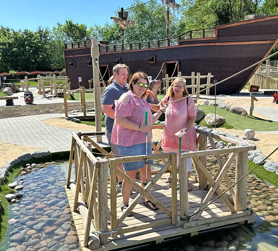 Three people play mini golf on wooden platform.