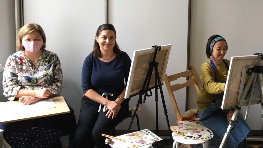Three women sitting, two painting on canvases.