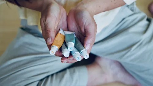 Hands holding tubes of paint, seated background.