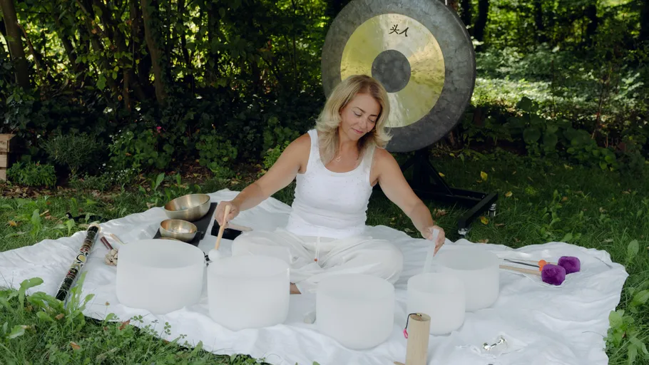 Woman plays crystal bowls outdoors on white mat.