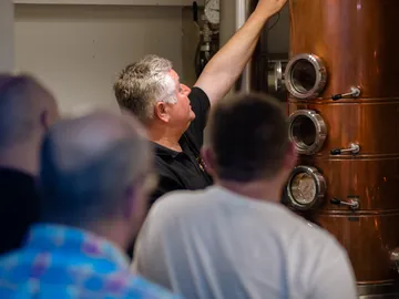 A man points to a copper distillery; visitors watch.