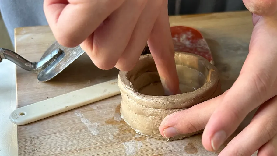 Hands shaping clay pot on wooden board.