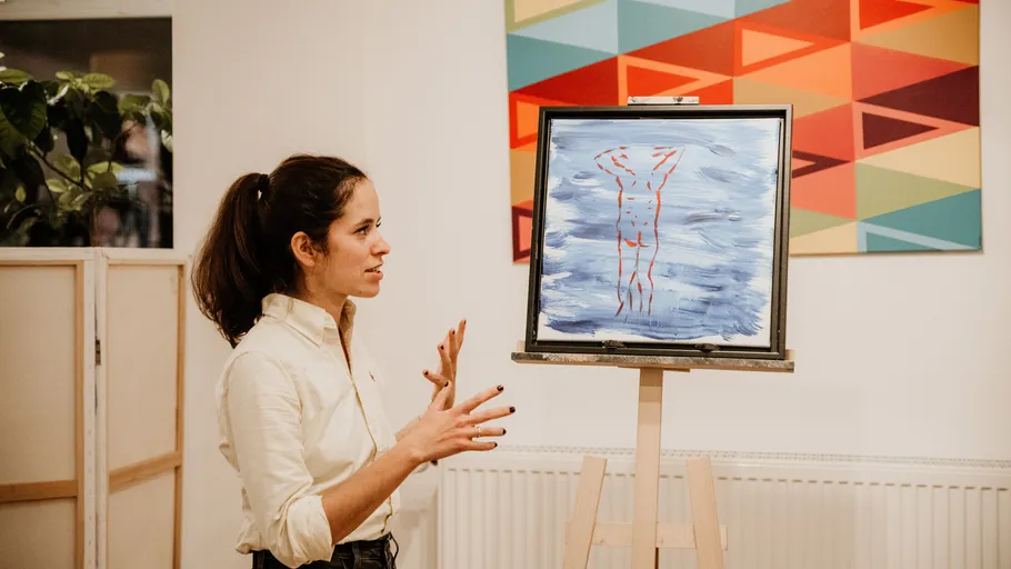 Woman explains abstract painting on easel indoors.