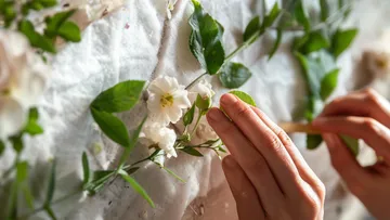Hände arrangieren Blumen und Blätter auf Stoff.