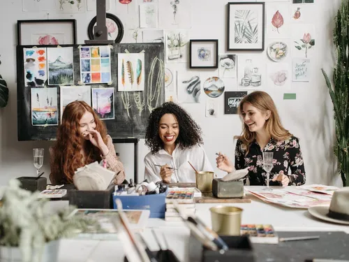 Drei Frauen sitzen lachend an einem Tisch in einem Atelier.