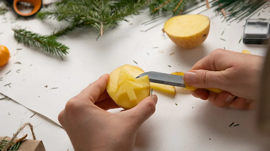 Hands carving potato with knife, surrounded by greenery.