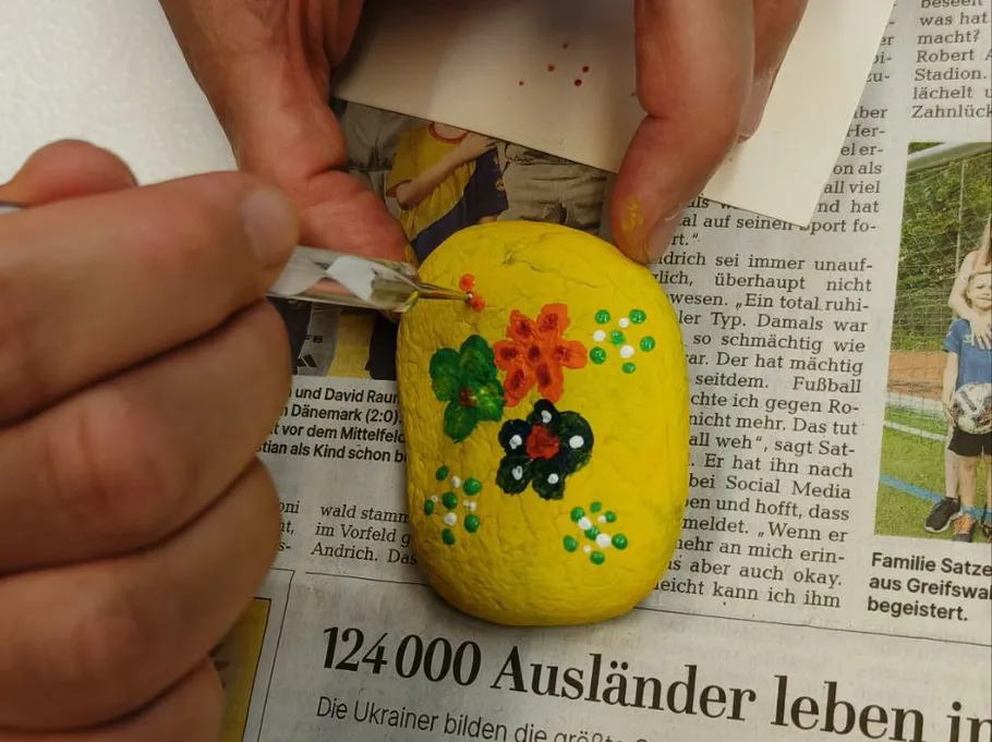 Hände bemalen gelben Stein mit bunten Blumen.