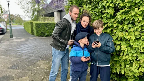 Familie schaut auf Handys nahe grüner Hecke.