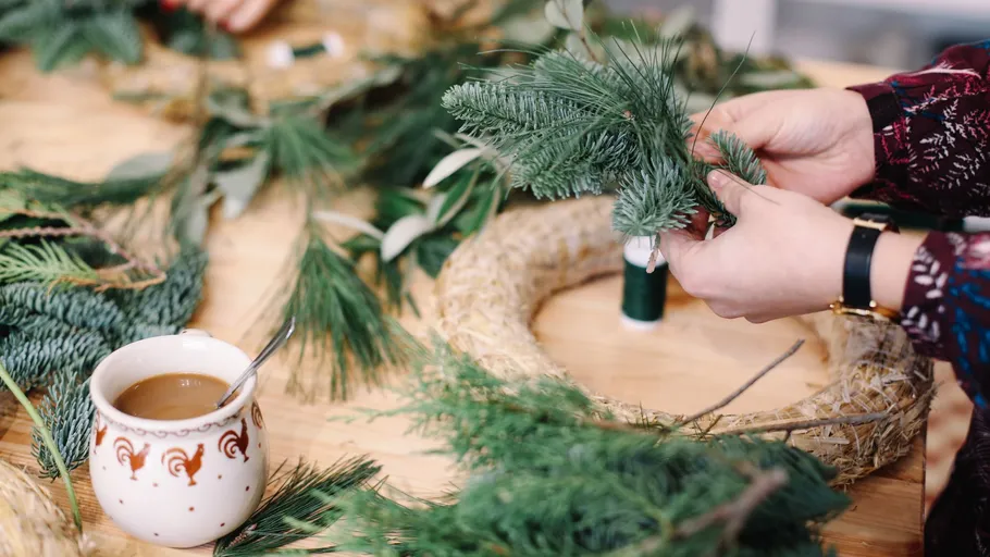 Hands crafting wreath with fir branches, cup nearby.