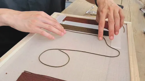 Hands arranging clay strands on wooden frame.