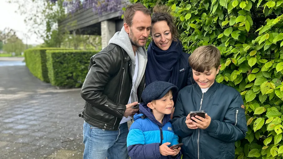 Family looking at phones near leafy hedge.