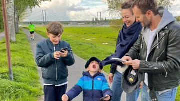 Family smiling with phones by riverside path.