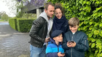 Familie schaut draußen auf Handys neben Büschen.