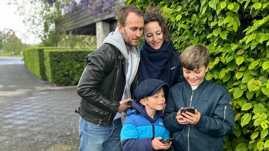 Family looking at smartphones near green hedge.